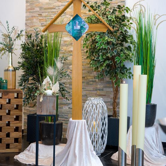 Holzkreuz mit Foto im Vordergrund, umgeben von Pflanzen und Kerzen auf einem eleganten Arrangement.