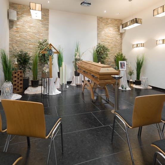 Trauerraum mit einem offenen holzernen Sarg, umgeben von Pflanzen, Kerzen und dekorativen Elementen.