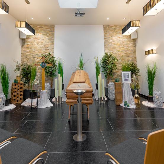 Ein Trauerraum mit einem Holzkoffer, Kerzen, Pflanzen und Stühlen zur Verfügung für Trauergäste.