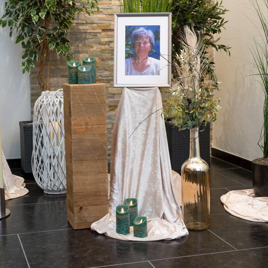 Ein gerahmtes Foto einer Frau steht auf einem Tisch, umgeben von Kerzen, Pflanzen und dekorativen Stoffen.