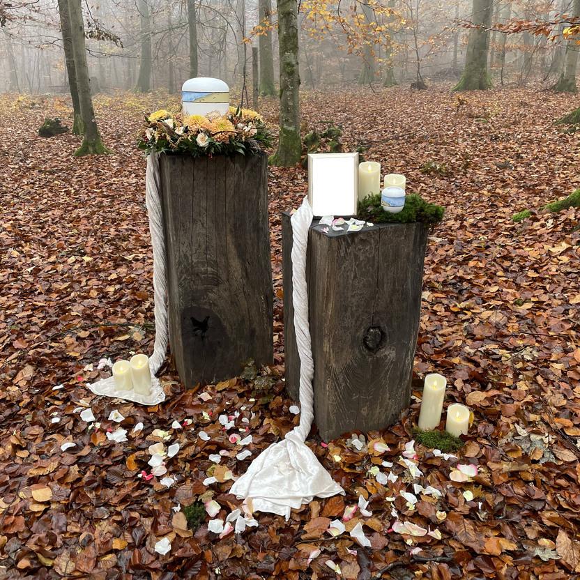 Zwei Holzskulpturen stehen im nebligen Wald, geschm&uuml;ckt mit Blumen, Kerzen und Bl&uuml;tenbl&auml;ttern auf dem Laub.