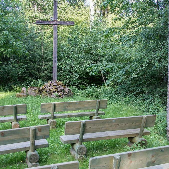 Gruppensitzgelegenheiten aus Holz um einen Steinhaufen mit Kreuz in einem schattigen Waldgebiet.