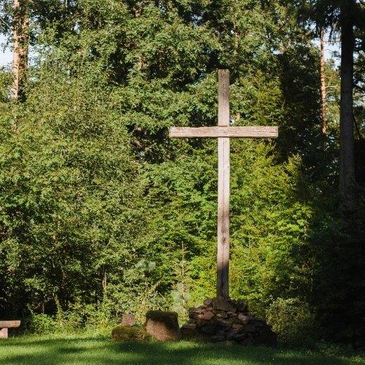 Holzkreuz auf einem Steinhaufen in einer Waldlichtung, umgeben von hohen B&auml;umen und einem Holzbank.