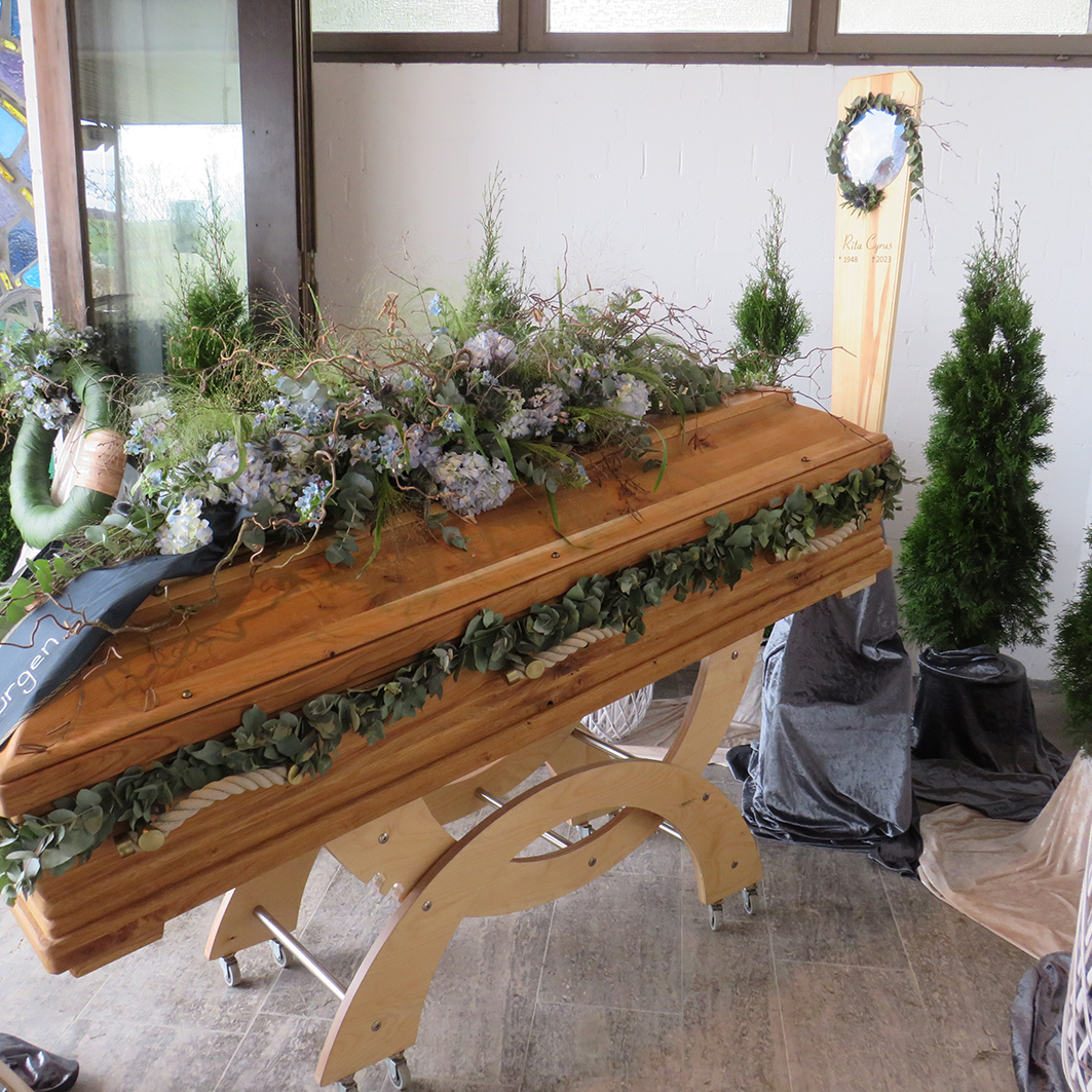 Holzurne mit floraler Dekoration und Trauerschleifen in einem Raum, umgeben von gr&uuml;nen Pflanzen und Tuch.