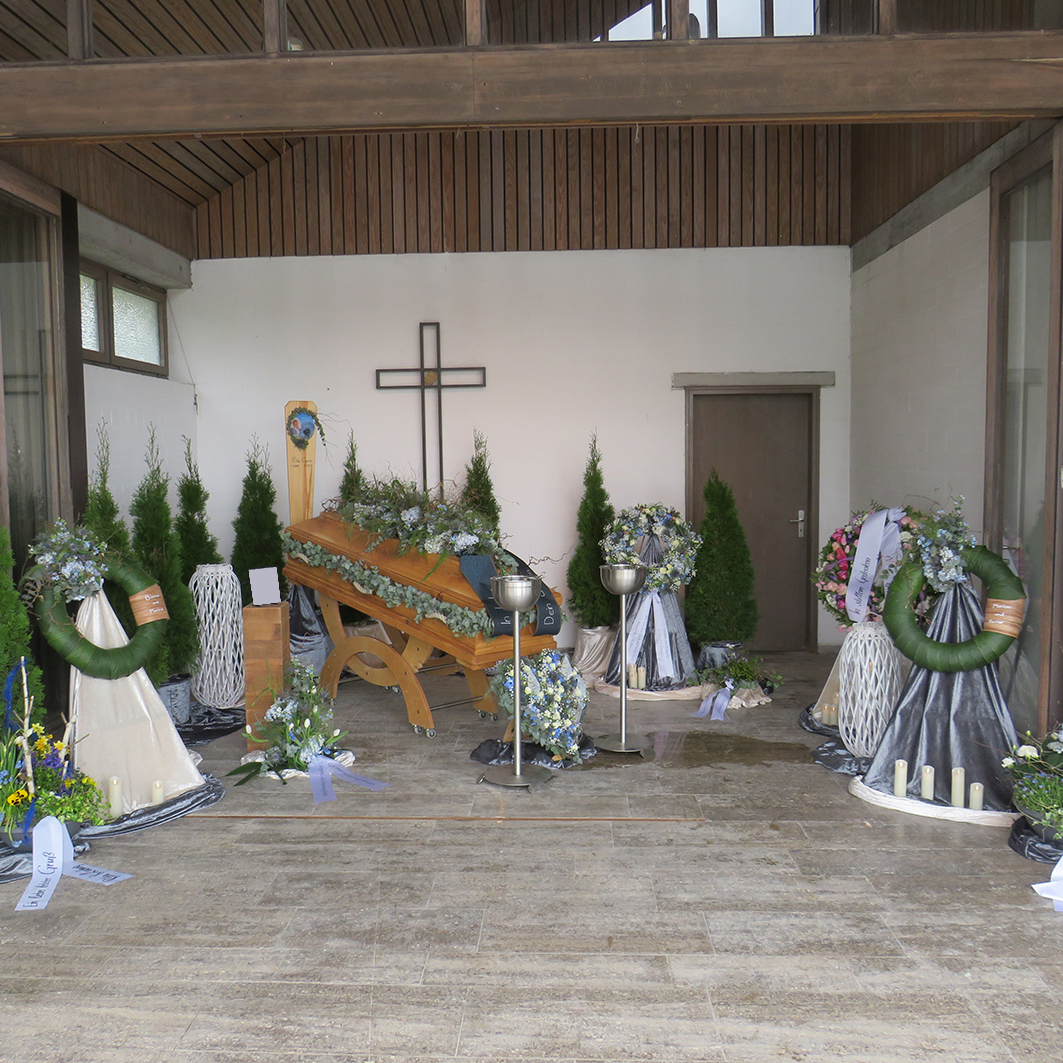 Andachtsraum mit einem Holzk&auml;fig und Trauerschmuck. Gr&uuml;ne Pflanzen und Blumenarrangements umgeben den Sarg.