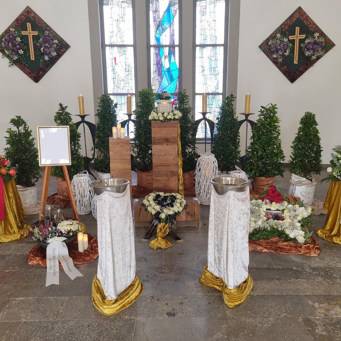 Blumengedeckte Trauerzeremonie mit Kerzen, einem Kreuz und einem leeren Bilderrahmen in einer chapelartigen Umgebung.