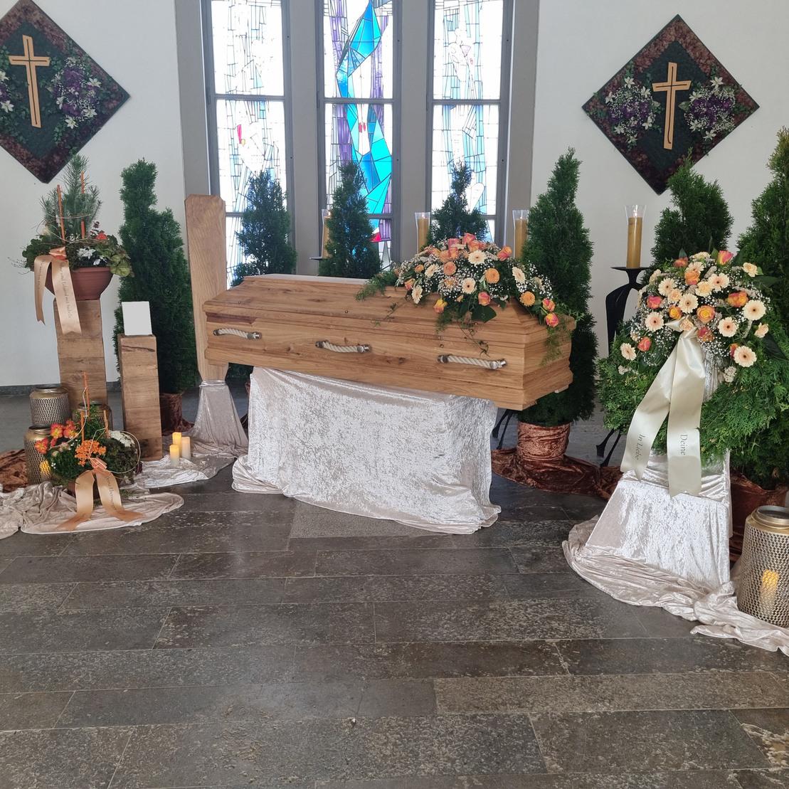 Dekorierte Trauerst&auml;tte mit einem Holzsarg, Blumenarrangements und Kerzen auf einem eleganten Tuch.