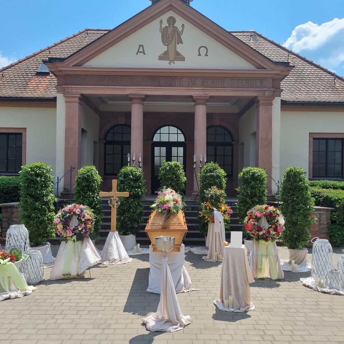 Feierliche Trauerzeremonie im Freien mit einem geschm&uuml;ckten Sarg, Blumenkr&auml;nzen und einer Kapelle im Hintergrund.
