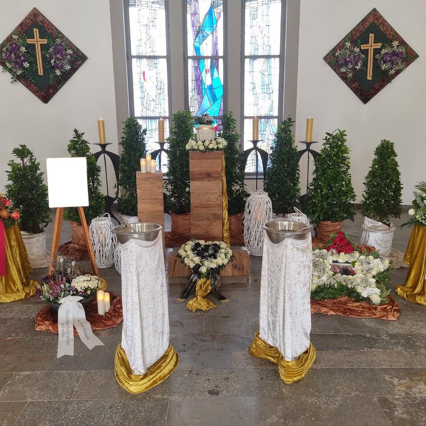 Dekorierter Raum mit Blumen, Kerzen und einem Kreuz, umgeben von B&auml;umen und einem Holzpodest f&uuml;r eine Trauerfeier.