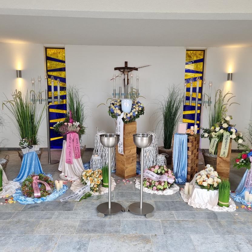 Dekorierte Ecke mit Blumenarrangements, Kerzen und Stoffen in verschiedenen Farben, sowie einem Kreuz im Hintergrund.