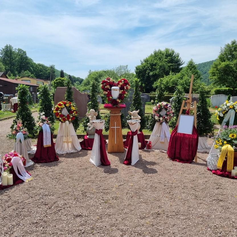 Dekorierte Grabstelle mit Blumenarrangements, Kerzen und einem Holzst&auml;nder auf einem Friedhof bei sonnigem Wetter.