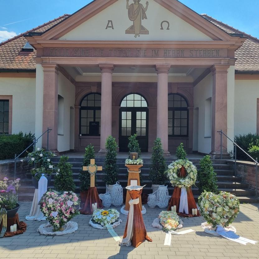 Blumenarrangements und Kr&auml;nze vor einer Kirche mit S&auml;ulen und einem Walmdach unter blauem Himmel.