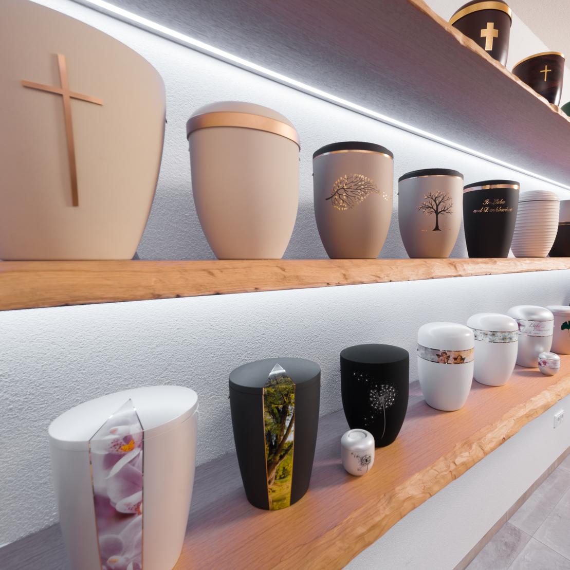 Reihe verschiedener Urnen mit dekorativen Motiven auf einem Holzregal, beleuchtet von einer LED-Leiste.