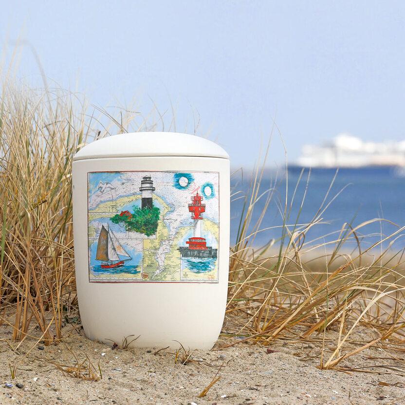 Müllbehälter mit maritimem Design steht im Sand, umgeben von Gräsern und Blick auf das Meer im Hintergrund.