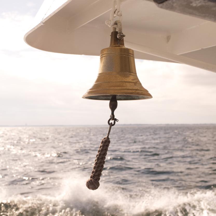 Goldene Schiffsglocke, aufgeh&auml;ngt an einem Boot, mit sch&auml;umenden Wellen im Hintergrund und einem bew&ouml;lkten Himmel.