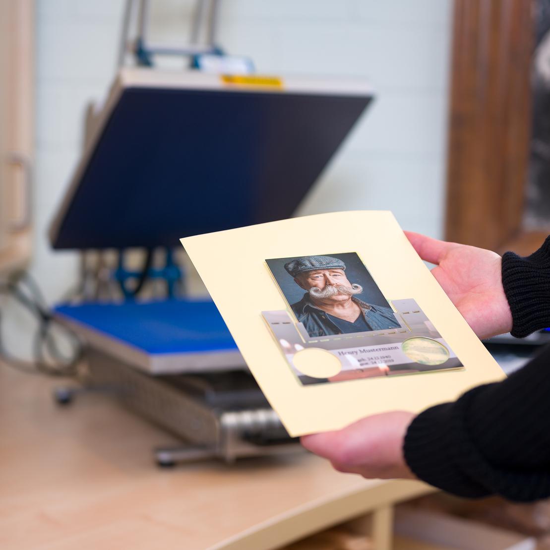 Person hält ein gerahmtes Porträt eines Mannes mit Schnurrbart und Mütze vor einer Druckmaschine auf einem Tisch.