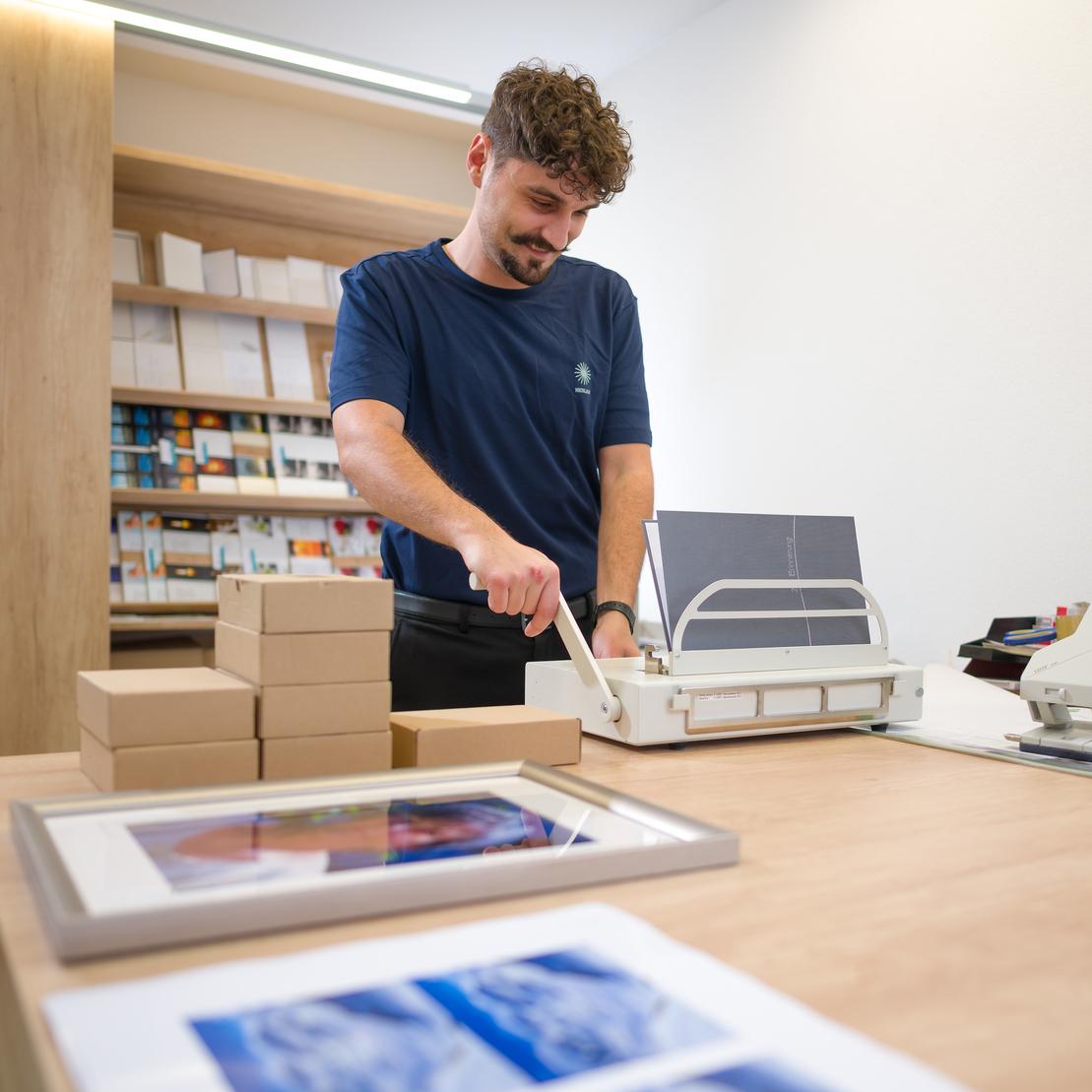 Mann schneidet Papiere an einem Tisch in einem Gesch&auml;ft, im Vordergrund sind gestapelte Kartons und Bilderrahmen zu sehen.