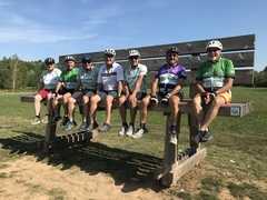Gruppe von sieben Radfahrern in Helmen sitzt auf einer Holzbank in einem Au&szlig;engebiet unter blauem Himmel.
