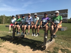 Gruppe von sieben Radfahrern in Helmen sitzt auf einer Holzbank in einem Außengebiet unter blauem Himmel.