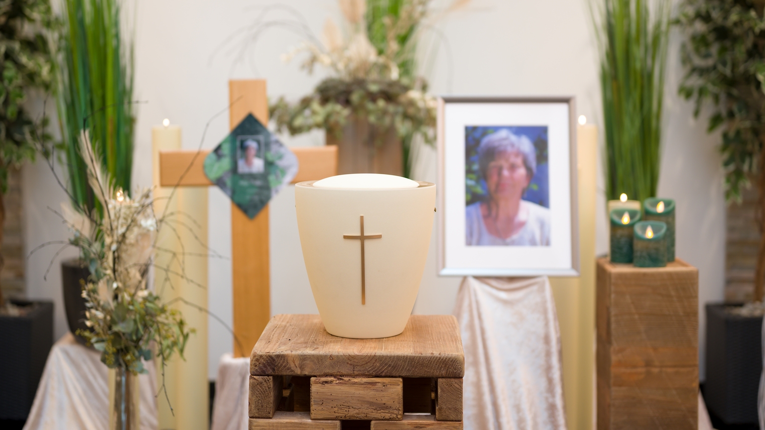 Auf einem Holzpodest steht eine Urne mit Kreuz, umgeben von Pflanzen, Kerzen und einem Bild einer Person.