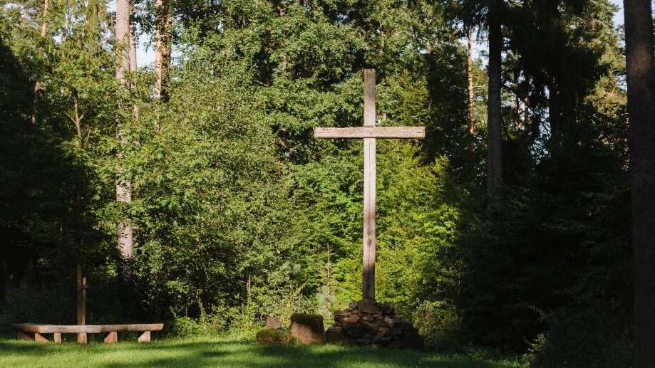 Holzkreuz, umgeben von B&auml;umen, mit einer steinernen Basis und einer Bank auf einer gr&uuml;nen Wiese im Wald.