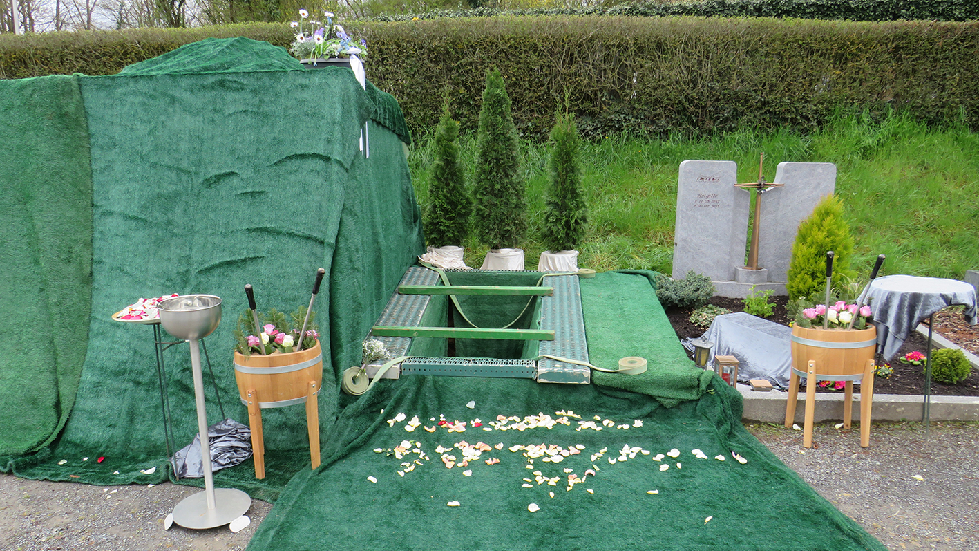 Begrünter Friedhof mit einer abgedeckten Grabanlage, zwei Blumengestecken und verschiedenen Trauerschmuckelementen.