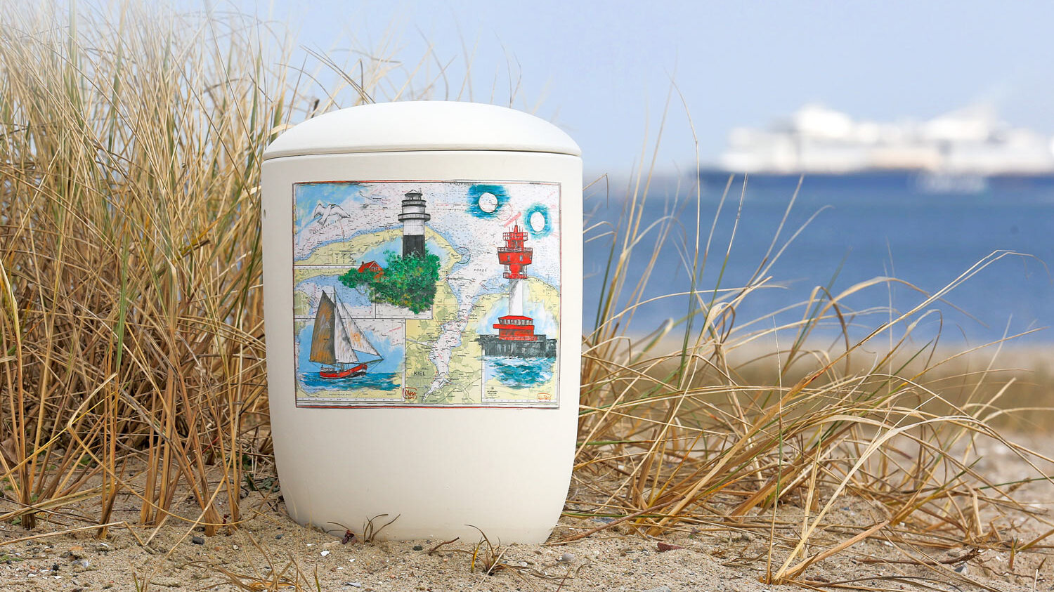 Abfallbeh&auml;lter mit maritimer Illustration steht im Sand vor einer K&uuml;stenlandschaft und Meer im Hintergrund.