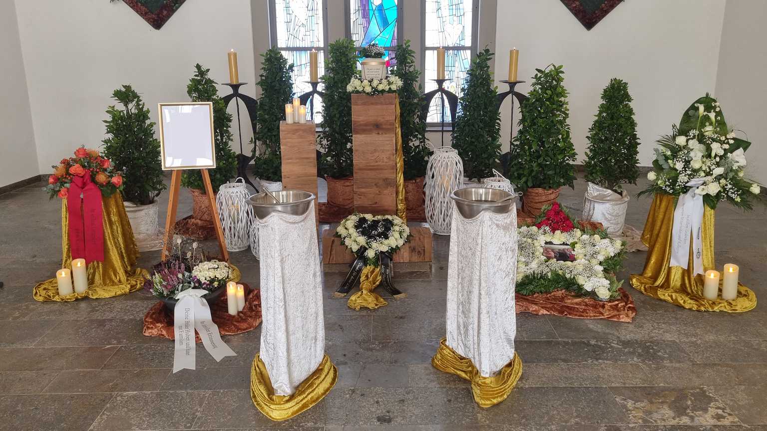 Dekorierte Trauerst&auml;tte mit Blumenarrangements, Kerzen, einem Kreuz und einem leeren Rahmen auf einem Holzpodest.
