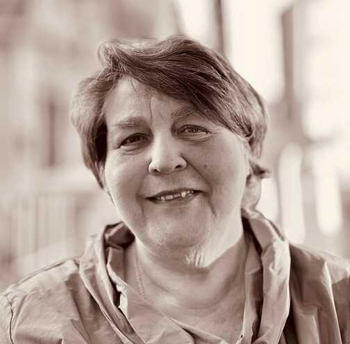 Portrait einer &auml;lteren Frau mit kurzer, lockiger Frisur, die freundlich in die Kamera l&auml;chelt. Hintergrund unscharf.