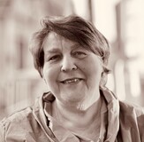 Portrait einer älteren Frau mit kurzer, lockiger Frisur, die freundlich in die Kamera lächelt. Hintergrund unscharf.