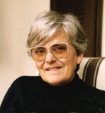 Porträt einer älteren Frau mit kurzen grauen Haaren, Brille und schwarzem Pullover, lächelnd vor einem gestreiften Hintergrund.