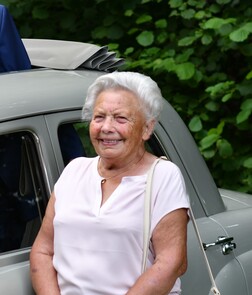 L&auml;chelnde &auml;ltere Frau mit grauem Haar in einem hellen Oberteil, neben einem klassischen Auto vor gr&uuml;ner Vegetation.