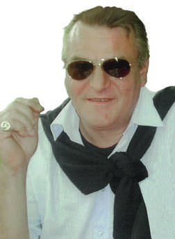 Mann mit Sonnenbrille und schwarzem Pullover um den Hals, l&auml;chelt und h&auml;lt eine Zigarette in der Hand.