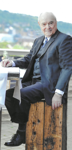 &Auml;lterer Mann in Anzug sitzt auf einem Holzblock, l&auml;chelt und schaut in die Kamera, mit einer Landschaft im Hintergrund.