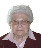 Porträt einer älteren Frau mit lockigem, grauem Haar, Brille und Perlenkette, in einem roten Oberteil vor weißem Hintergrund.