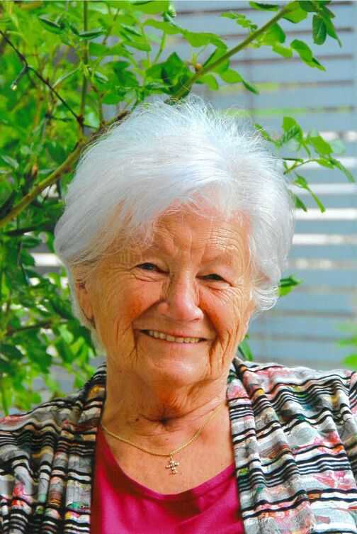 L&auml;chelnde &auml;ltere Frau mit grauen Haaren, in bunter Jacke, sitzt vor einer gr&uuml;nen Pflanze.