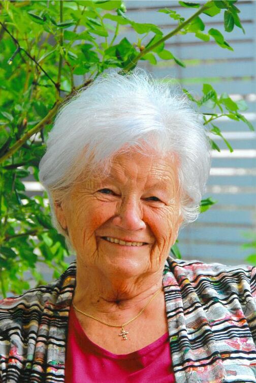 L&auml;chelnde &auml;ltere Frau mit grauen Haaren, in bunter Jacke, sitzt vor einer gr&uuml;nen Pflanze.