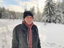 Älterer Mann mit Mütze und Schal steht lächelnd im Schnee vor verschneitem Wald.