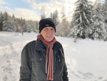 Älterer Mann mit Mütze und Schal steht lächelnd im Schnee vor verschneitem Wald.