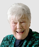 Lächelnde ältere Frau mit kurzen grauen Haaren, trägt einen grünen, gemusterten Schal vor einem hellen Hintergrund.