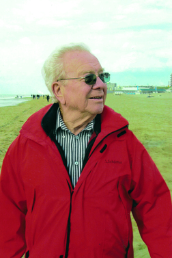 &Auml;lterer Mann mit Brille und roter Jacke, der am Strand l&auml;chelt und auf das Meer schaut. Hintergrund mit leicht bew&ouml;lktem Himmel.