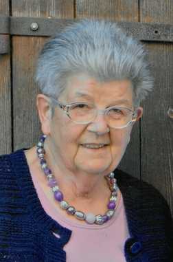 &Auml;ltere Frau mit grauen Haaren, Brille und lila Perlenkette, steht vor einer Holzpaneelwand und l&auml;chelt freundlich.
