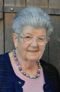 &Auml;ltere Frau mit grauen Haaren, Brille und lila Perlenkette, steht vor einer Holzpaneelwand und l&auml;chelt freundlich.