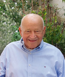 Lächelnder älterer Mann in hellblauer Hemdbluse vor einer blühenden Gartenlandschaft.