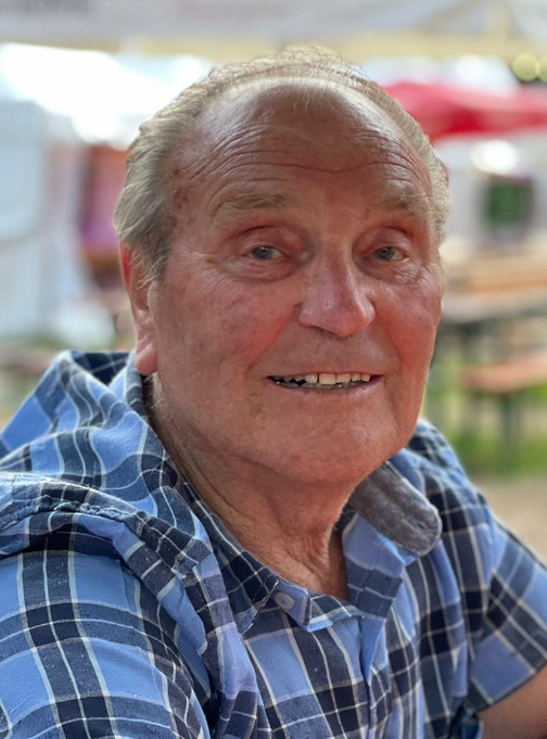 &Auml;lterer Mann mit l&auml;chelndem Gesicht, tr&auml;gt ein kariertes Hemd, sitzt im Freien mit unscharfem Hintergrund.