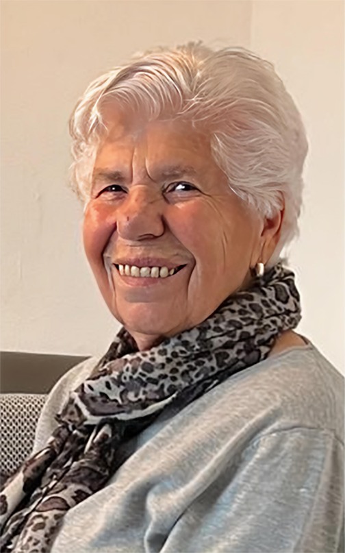 L&auml;chelnde &auml;ltere Frau mit wei&szlig;em Haar und einem tiermotivierten Schal sitzt vor einer hellen Wand.