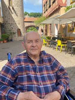 &Auml;lterer Mann in karierter Jacke sitzt im Rollstuhl in einer von Geb&auml;uden umgebenen Gasse mit Freisitz.
