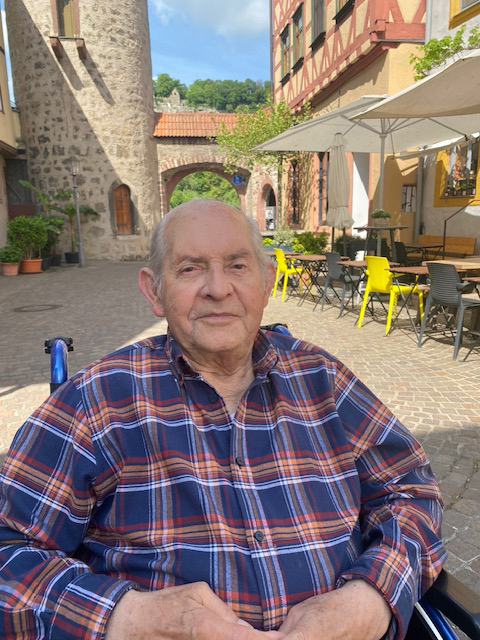 &Auml;lterer Mann in karierter Jacke sitzt im Rollstuhl in einer von Geb&auml;uden umgebenen Gasse mit Freisitz.