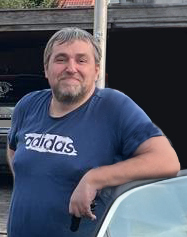 Mann mit kurzen Haaren in blauem T-Shirt lehnt l&auml;chelnd an einem Auto vor einer Garage.