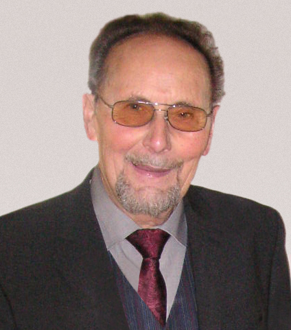 Portr&auml;t eines l&auml;chelnden &auml;lteren Mannes mit Brille, in schwarzem Anzug und rot-schwarzer Krawatte. Neutrale Hintergrundfarbe.